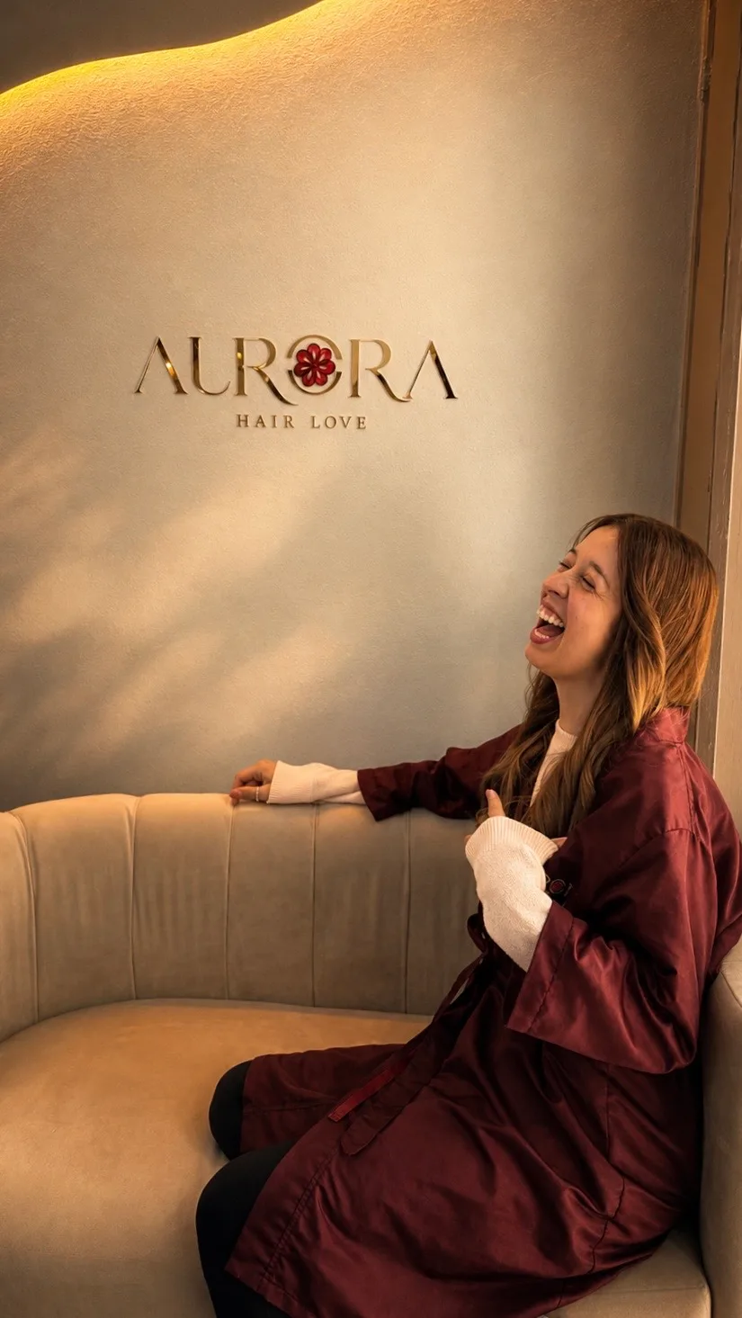 El logotipo dorado de Aurora Hair Love en la pared, con una clienta riendo en el sofá.